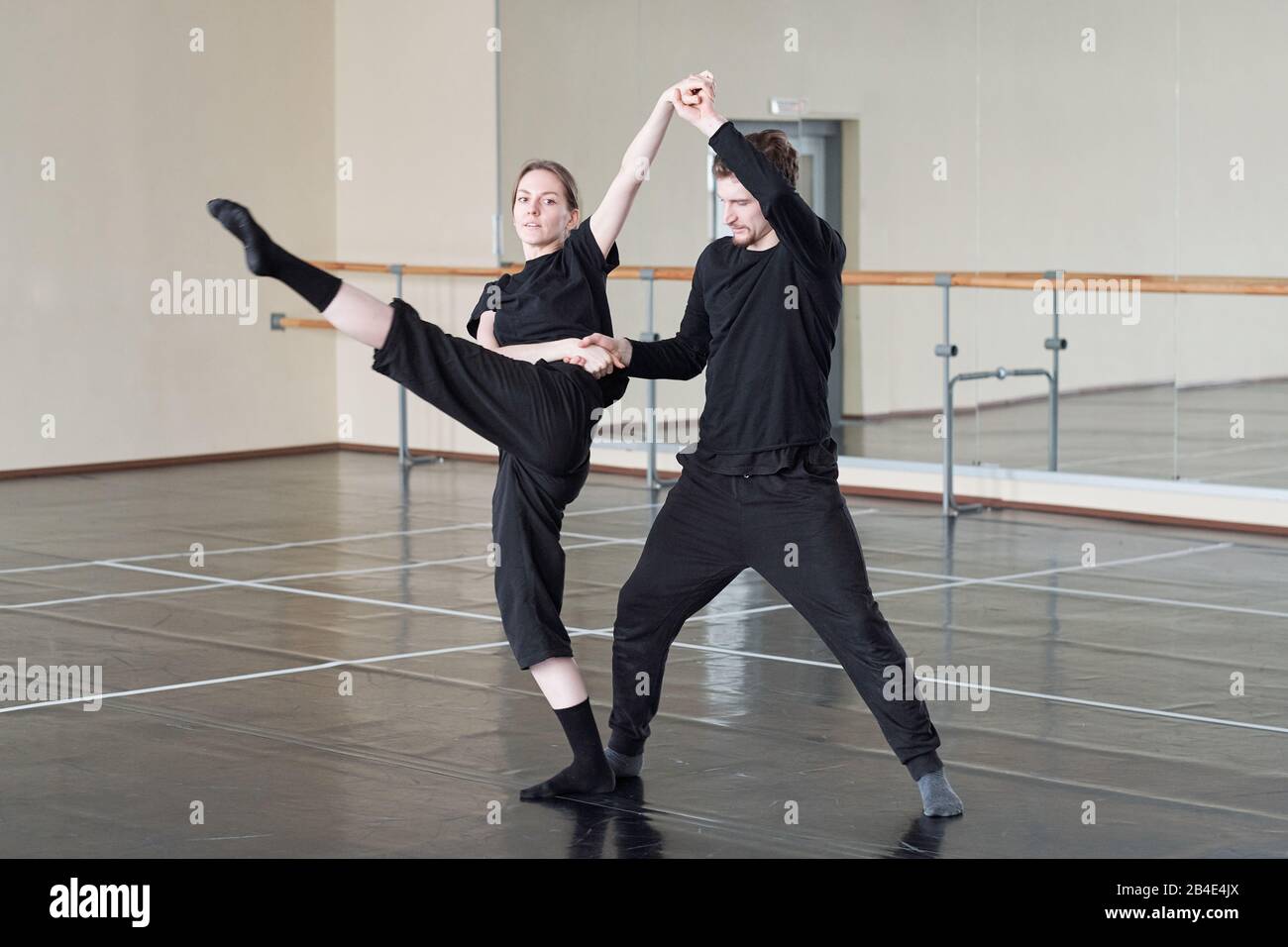 Young woman stretching one leg forward while holding by hands with her trainer during dance exercise in large studio Stock Photo
