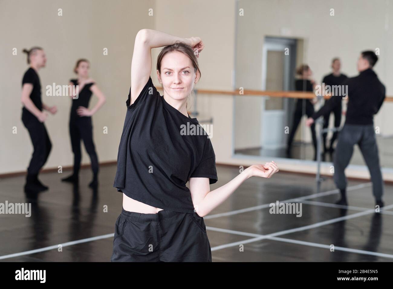 Pretty young student of modern ballet dancing course in black activewear standing in front of camera while doing exercise Stock Photo
