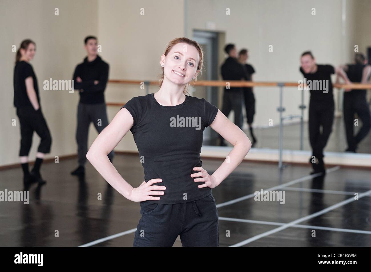 Young cheerful instructor of modern dancing course standing in akimbo pose in front of camera with students on background Stock Photo