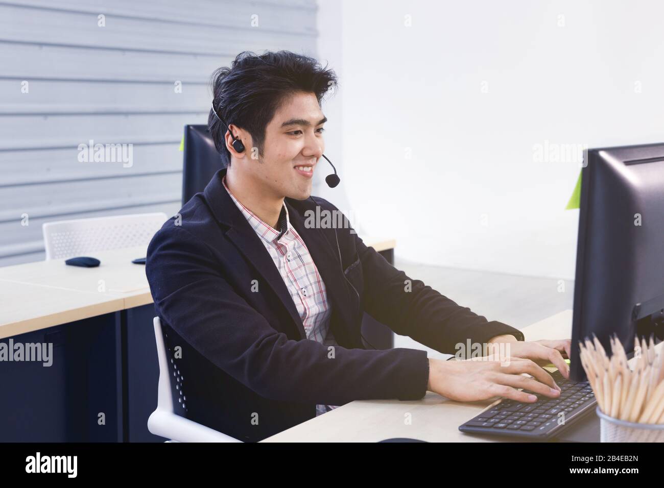 Smiling handsome Asian call center operator use computer for communicating with customer or perform other assignment. Stock Photo