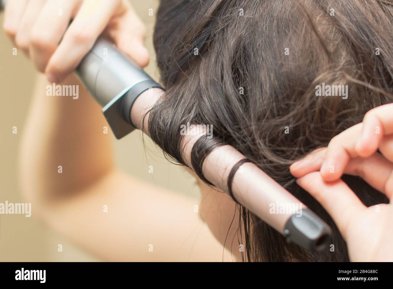 Hairdresser is making a curly hair. A hair styling using a ceramic curling wand waver. Young woman is styling her brown straight hair Stock Photo