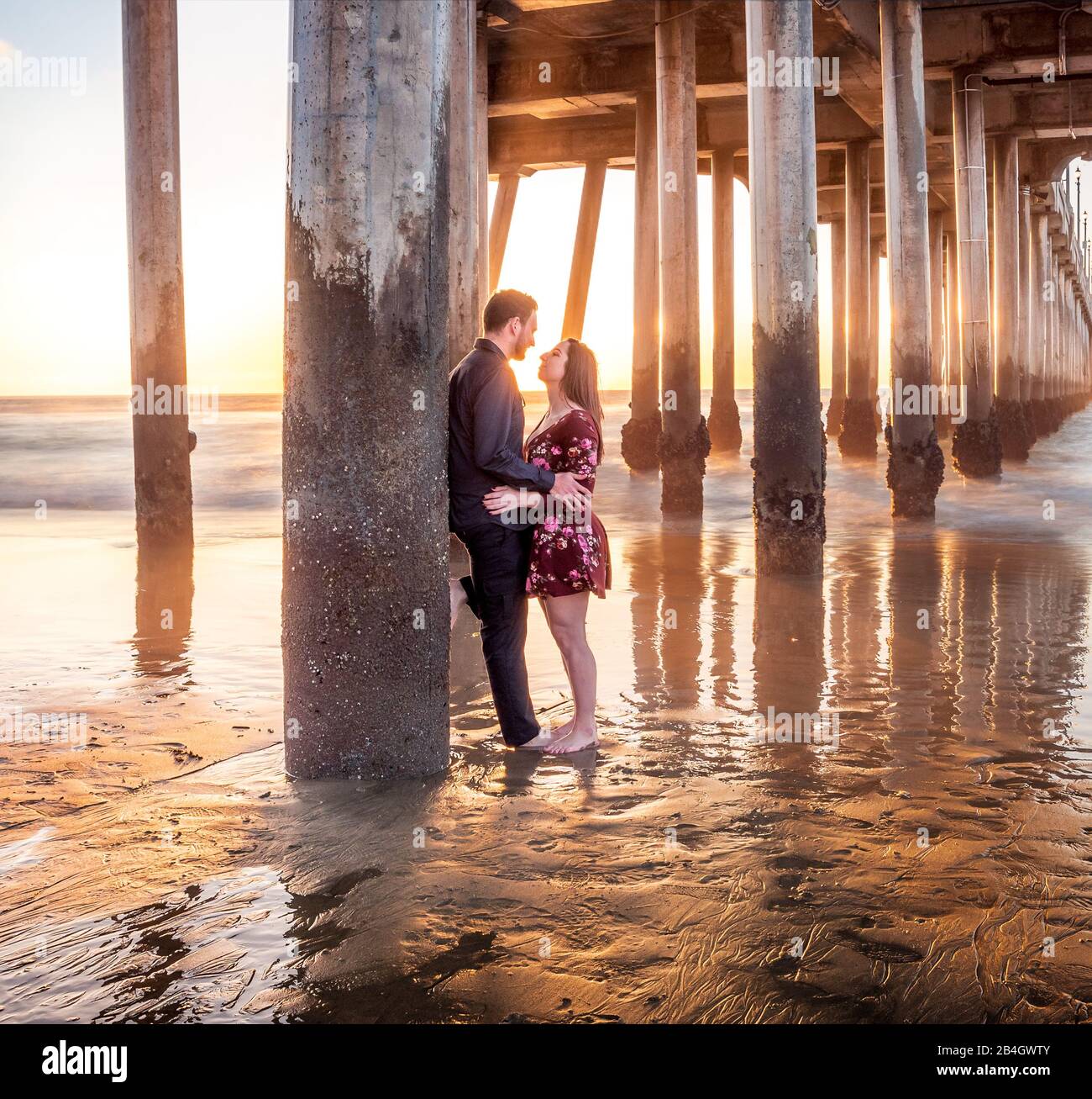 A romantic couple embracing below a pier on the beach at sunset Stock Photo