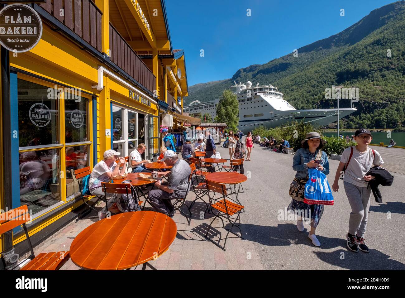 Tourists in Flåm, FACTORY KUTSALG FACTORY OUTLET, café, yellow wooden house, tour boat, fjord, mountain, trees, blue sky, Sogn og Fjordane, Norway, Scandinavia, Europe Stock Photo