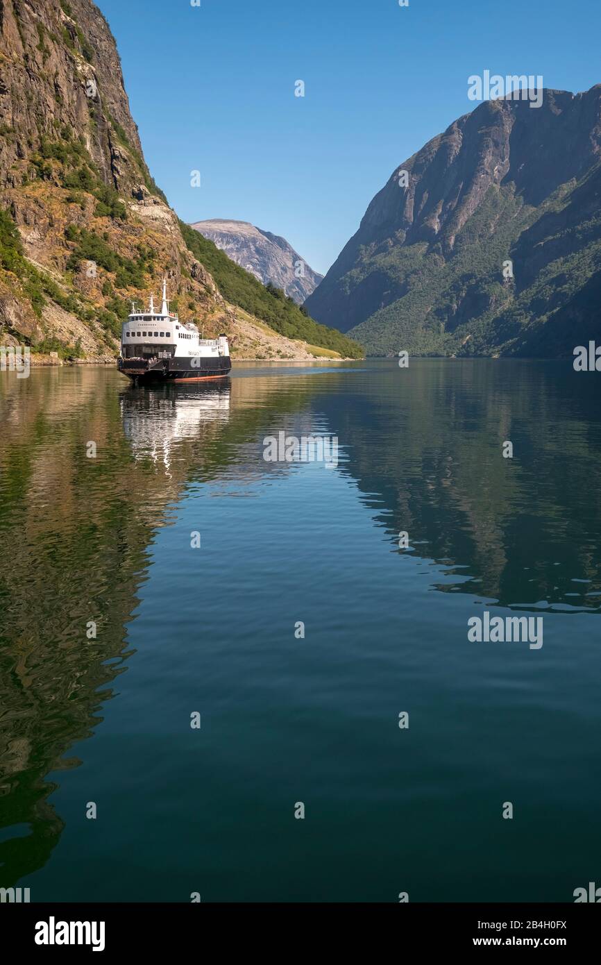 A sailing cruise ship on a beautiful fjord in which reflects the light, surrounded by rocks that are partly overgrown with trees, above it a bright blue sky. Gudvangen, Sogn og Fjordane, Norway, Scandinavia, Europe Stock Photo