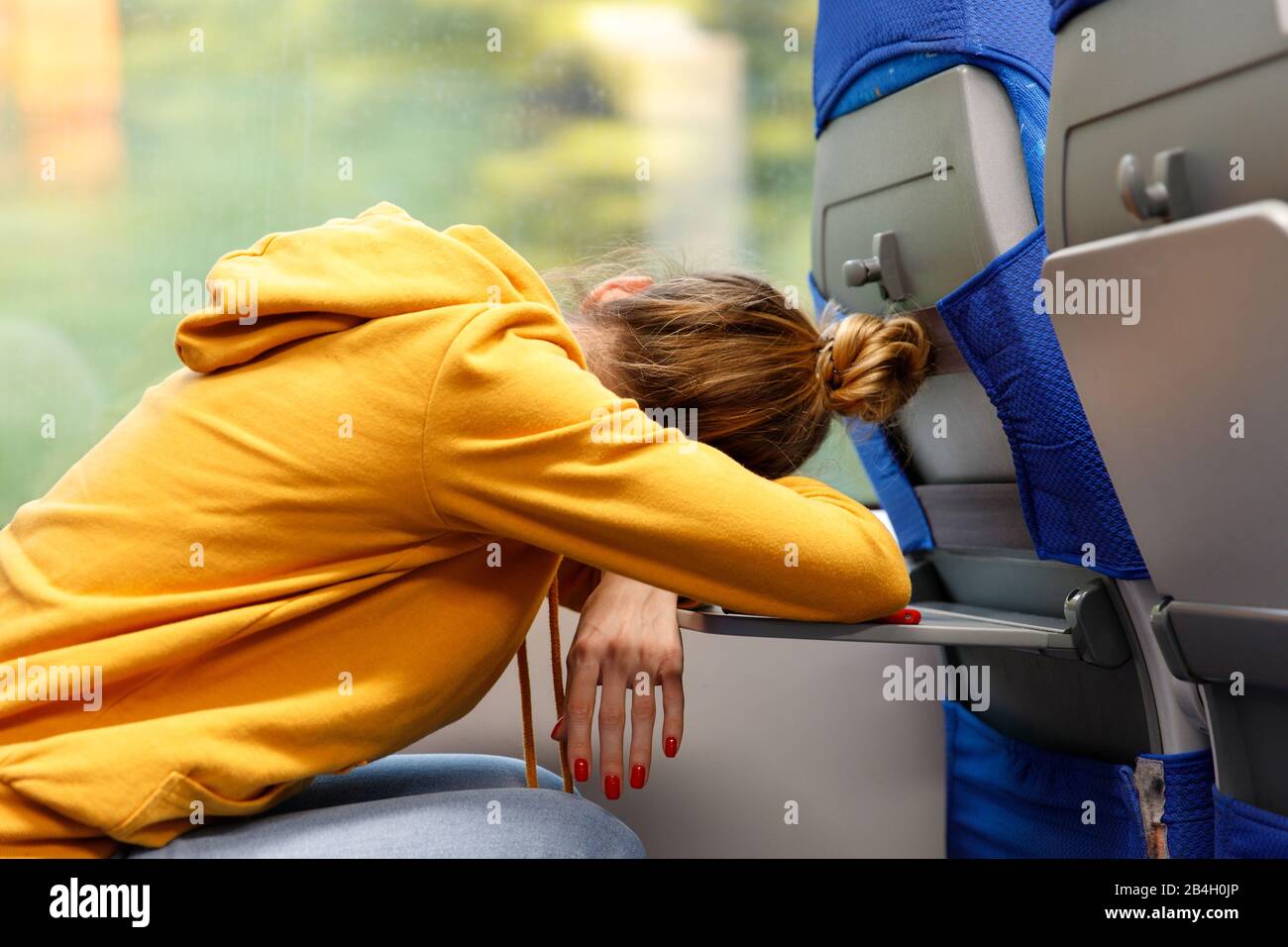 Woman in orange hoodie sitting in a seat and sleeping on a folding table in public transportation. Tired exhausted young female after work asleep in t Stock Photo