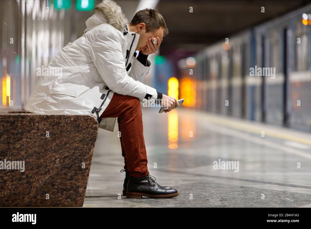 Crying man in white parka very upset, holding smartphone, gets bad news, covers his face with his hand, sitting on bench in subway station. Problem in Stock Photo