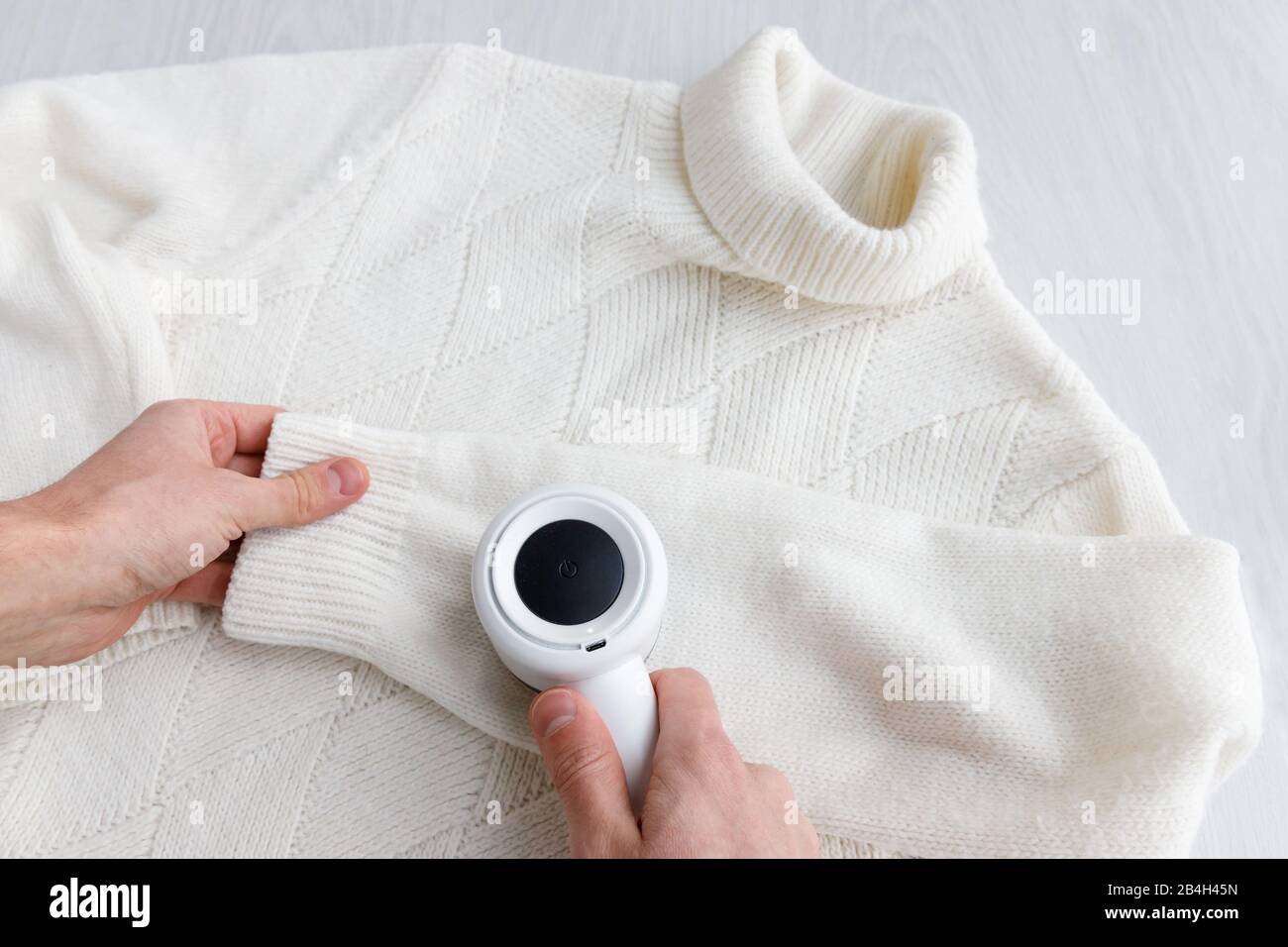 Man's hand holding a lint remover from a white acrylic or wool sweater. Electric device after cleaning and collected fluff/lint, light wooden backgrou Stock Photo