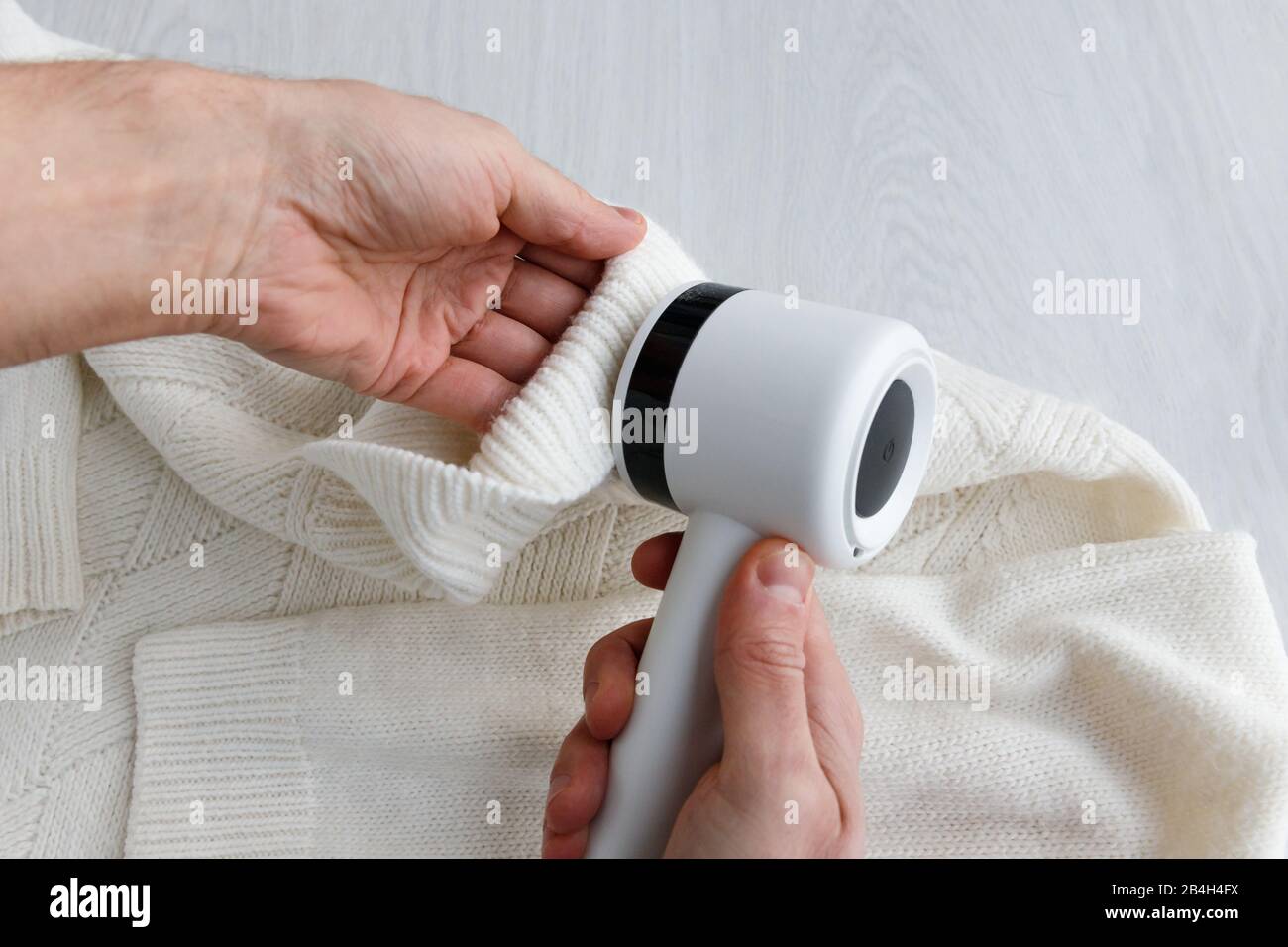 Man's hand holding a lint remover from a white acrylic or wool sweater. Electric device after cleaning and collected fluff/lint, light wooden backgrou Stock Photo