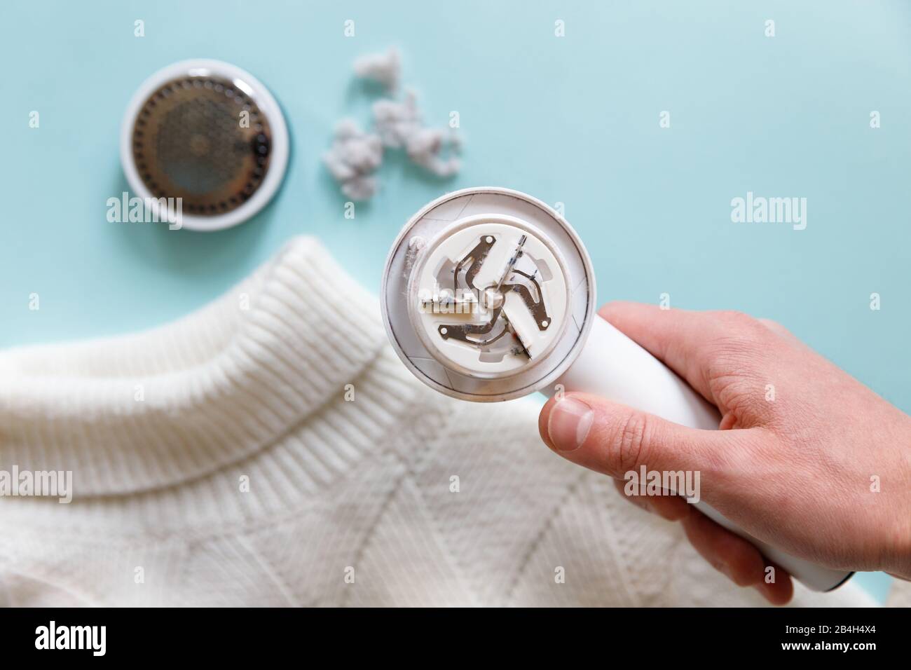 Man's hand holding a lint remover from a white acrylic or wool sweater. Electric device after cleaning and collected fluff/lint, light wooden backgrou Stock Photo