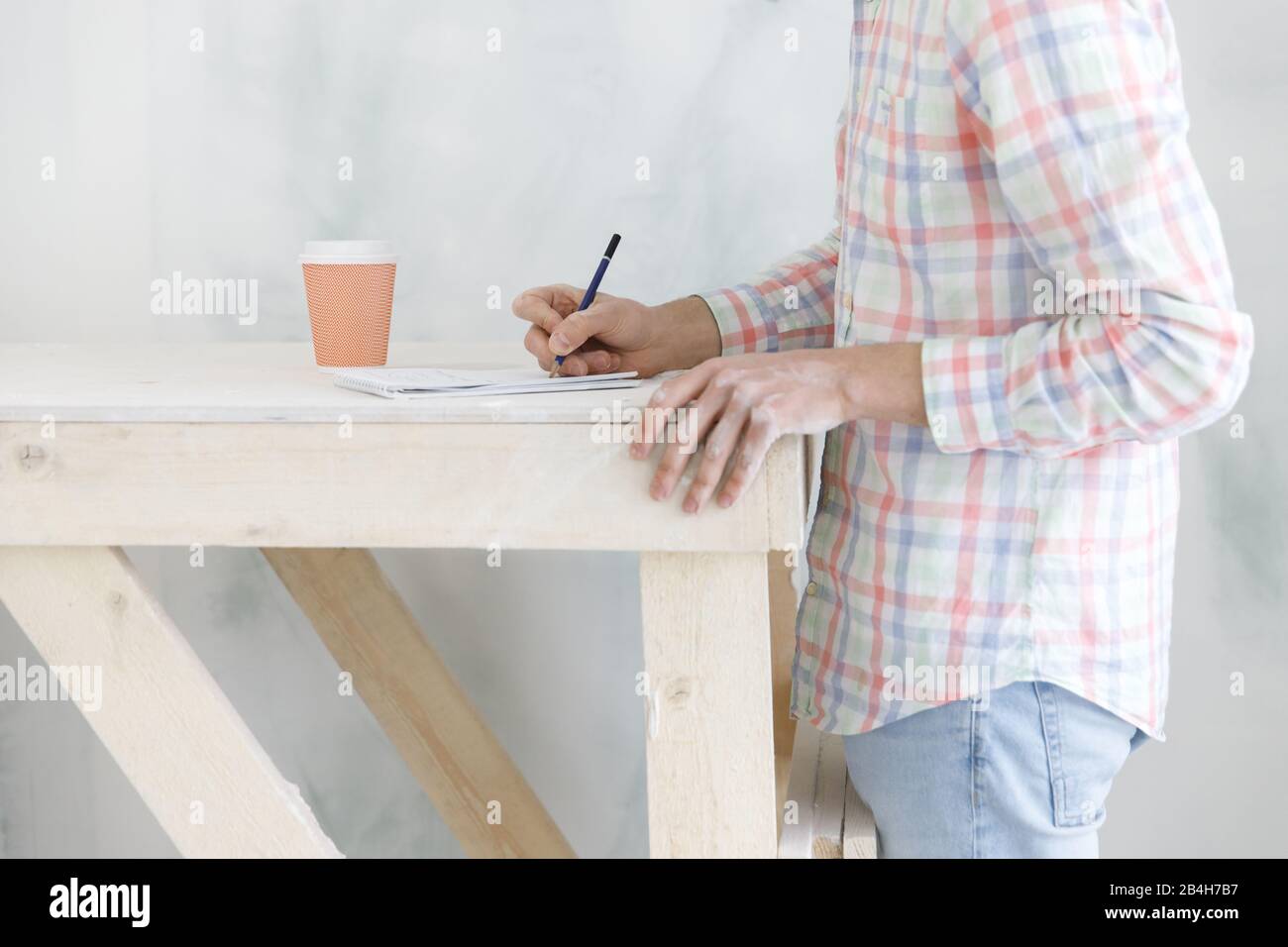 Cropped image of man in checked shirt, blue jeans having a coffee break from renovating, writes upcoming purchases in his notebook, soft focus. Home r Stock Photo