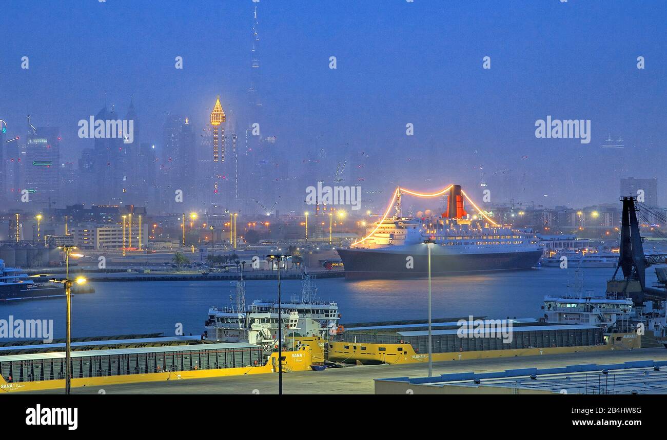 Hotel and museum ship Queen Elizabeth 2 (QE2) in the harbor with city skyline and Burj Khalifa 828m at night, Dubai, Persian Gulf, United Arab Emirates Stock Photo