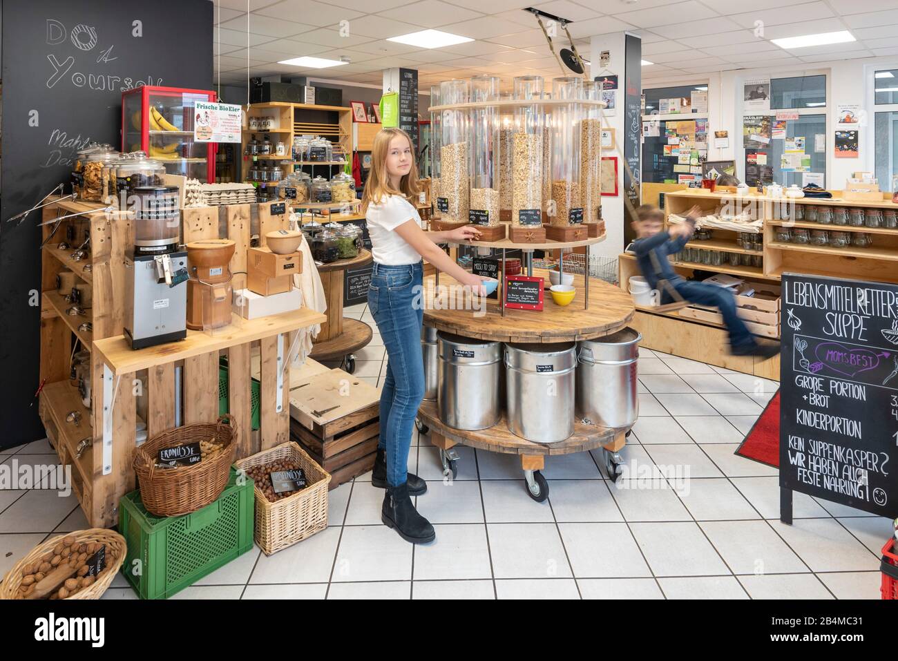 Eine junge Kundin steht vor einem Regal mit Abfüllbehältern für Müsli in einem Unverpacktladen. Rechts daneben schaukelt ein Junge. Szene aus einem Un Stock Photo