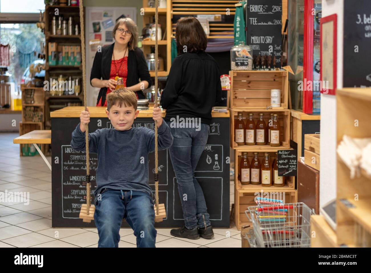 Ein Junge schaukelt in einem Unverpacktladen. Im Hintergrund berät die Besitzerin eine Kundin. Stock Photo