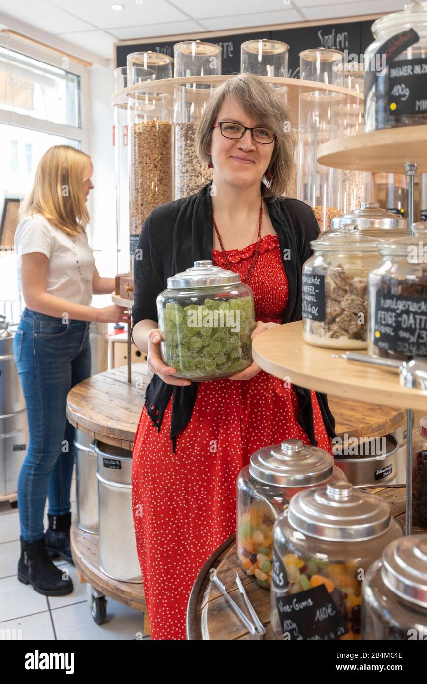 Eine Frau hält in einem Unverpacktladen ein Glas mit Süßigkeiten in den Händen. Im Hintergrund steht eine junge Kundin an einem Regal mit Abfüllbehält Stock Photo
