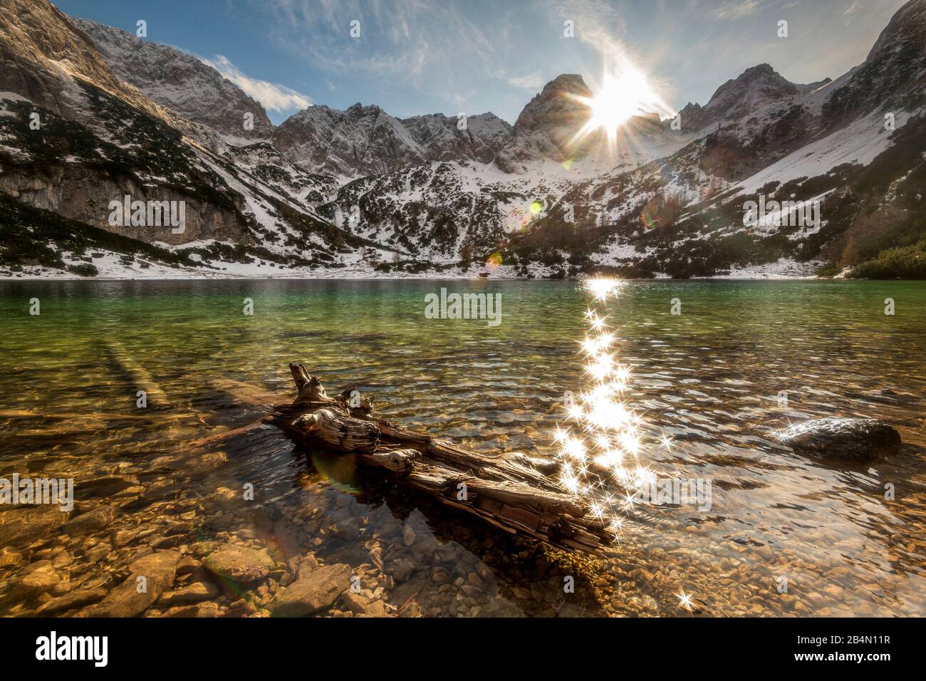 Sun rays are reflected in the Seebensee. An old log on the bank points to the clear, turquoise water in winter. Stock Photo