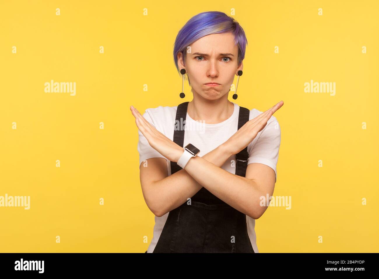 Portrait of serious hipster girl with violet short hair in denim overalls making x sign with crossed hands meaning definite no, never again, warning g Stock Photo