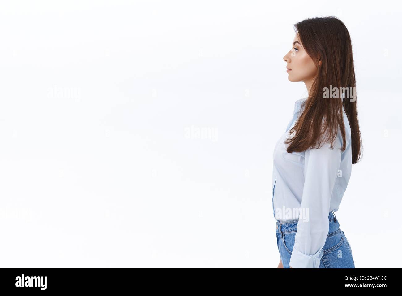 Profile portrait of skinny attractive brunette caucasian adult woman in blue shirt and jeans, standing straight and looking left at blank white space Stock Photo