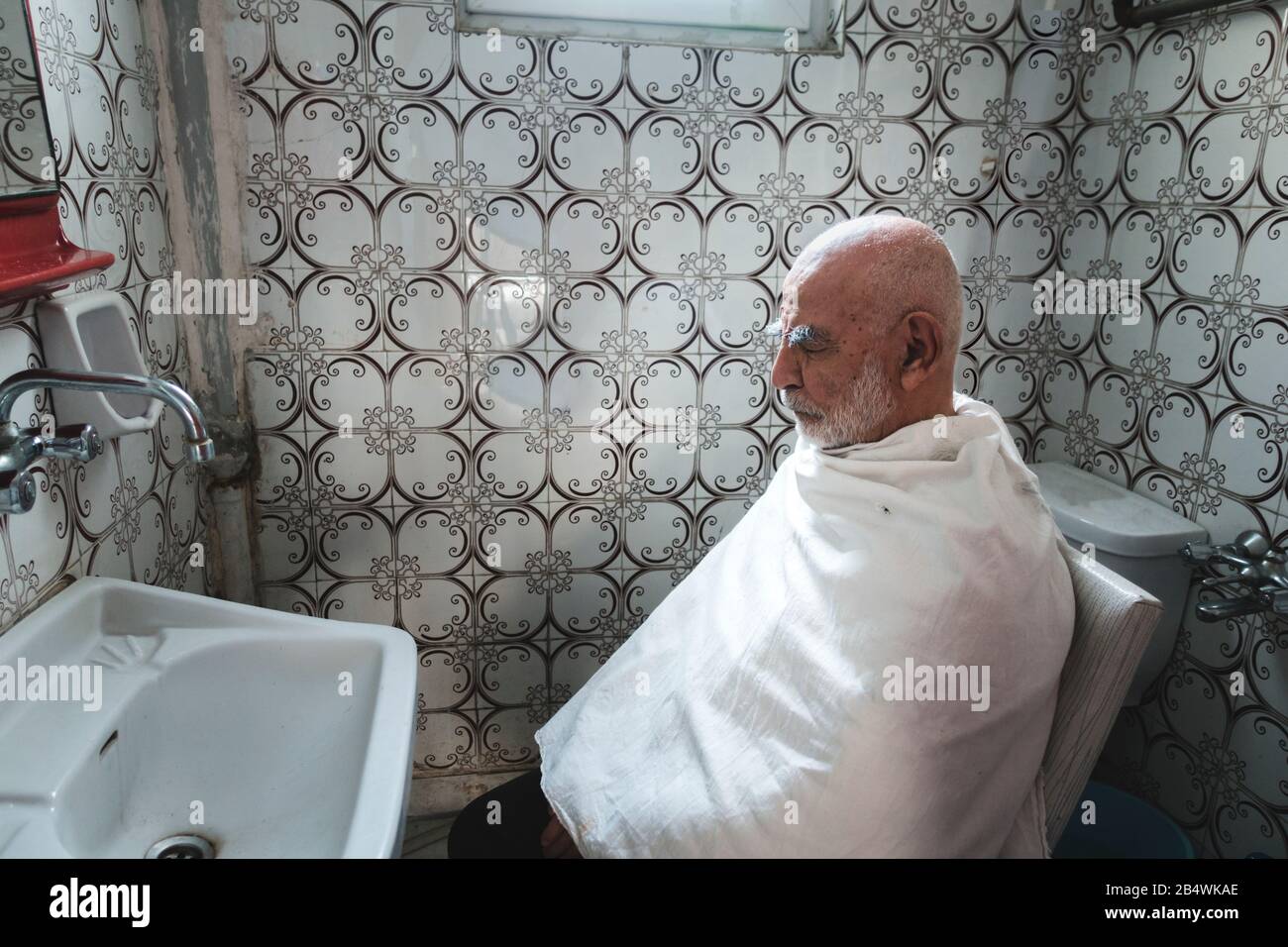 Very Old, White Haired, Bald Man waits for his Haircut In Old Vintage retro style tiled Bathroom Stock Photo