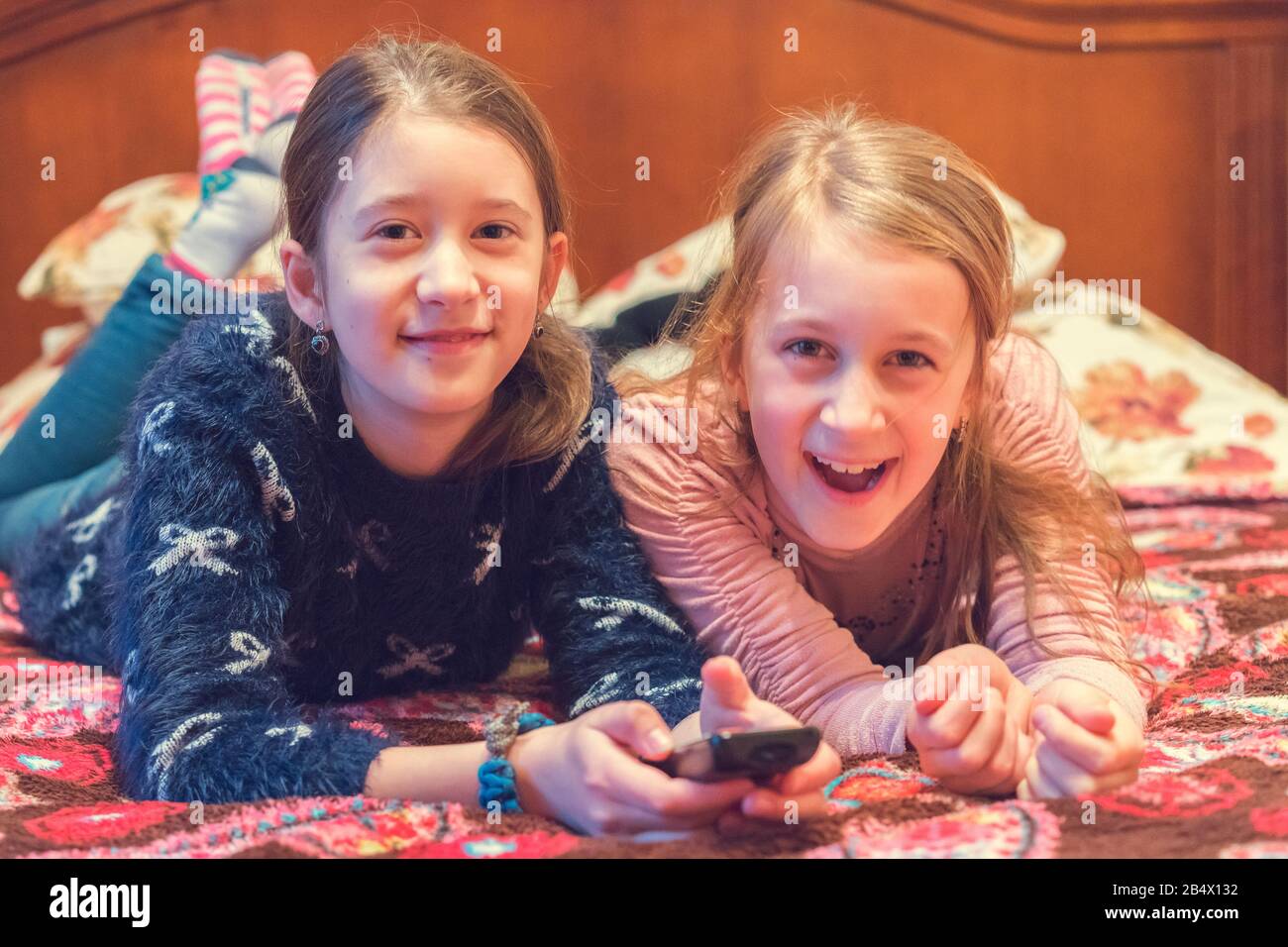 Two happy girls watching tv or movie. A horizontal photo of two young pretty smiling sisters lying on bed and watching their favourite serials. Stock Photo