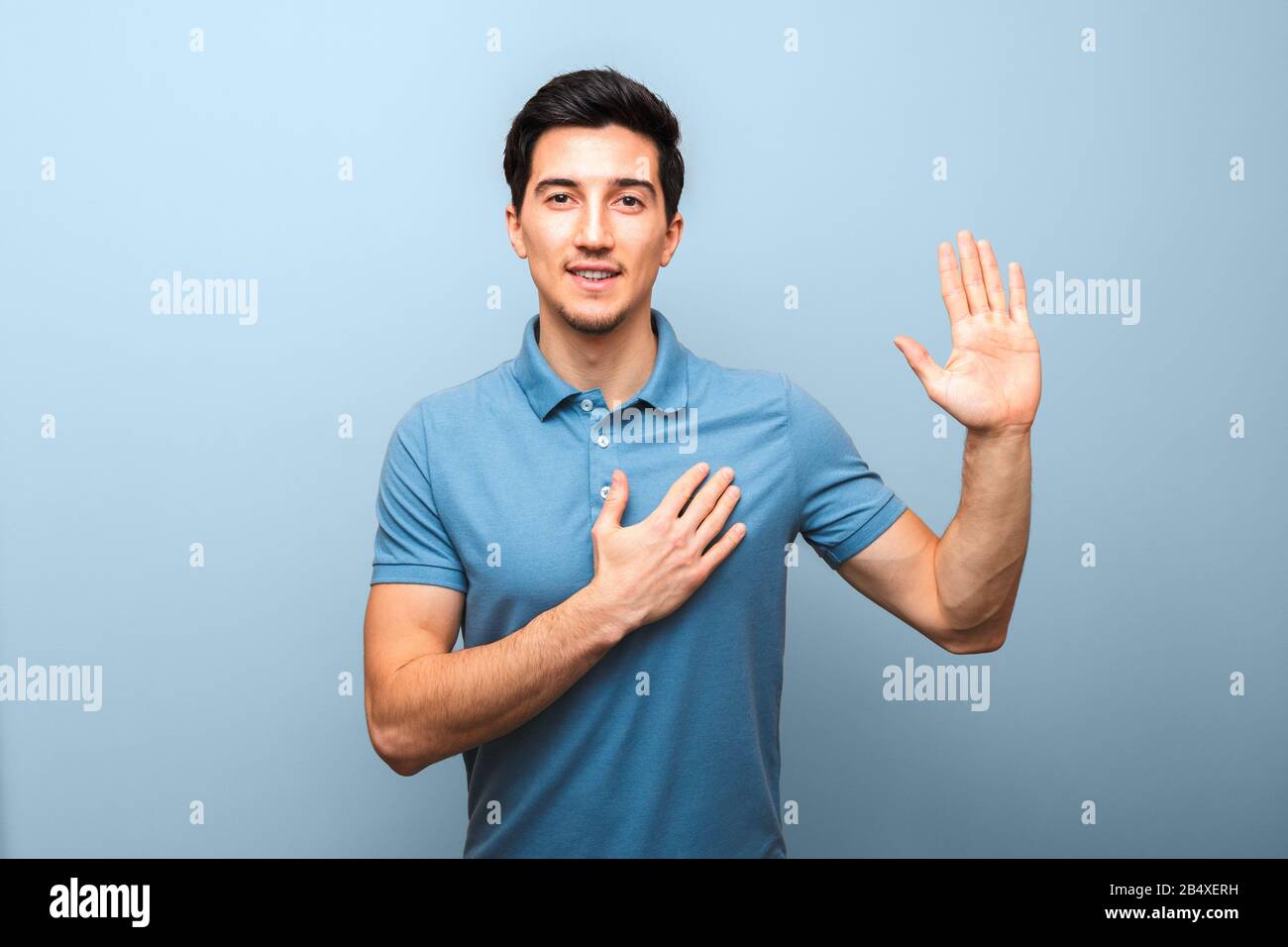 handsome young man with neutral smile in blue polo shirt with hand on chest giving oath against blue background. studio shoot Stock Photo