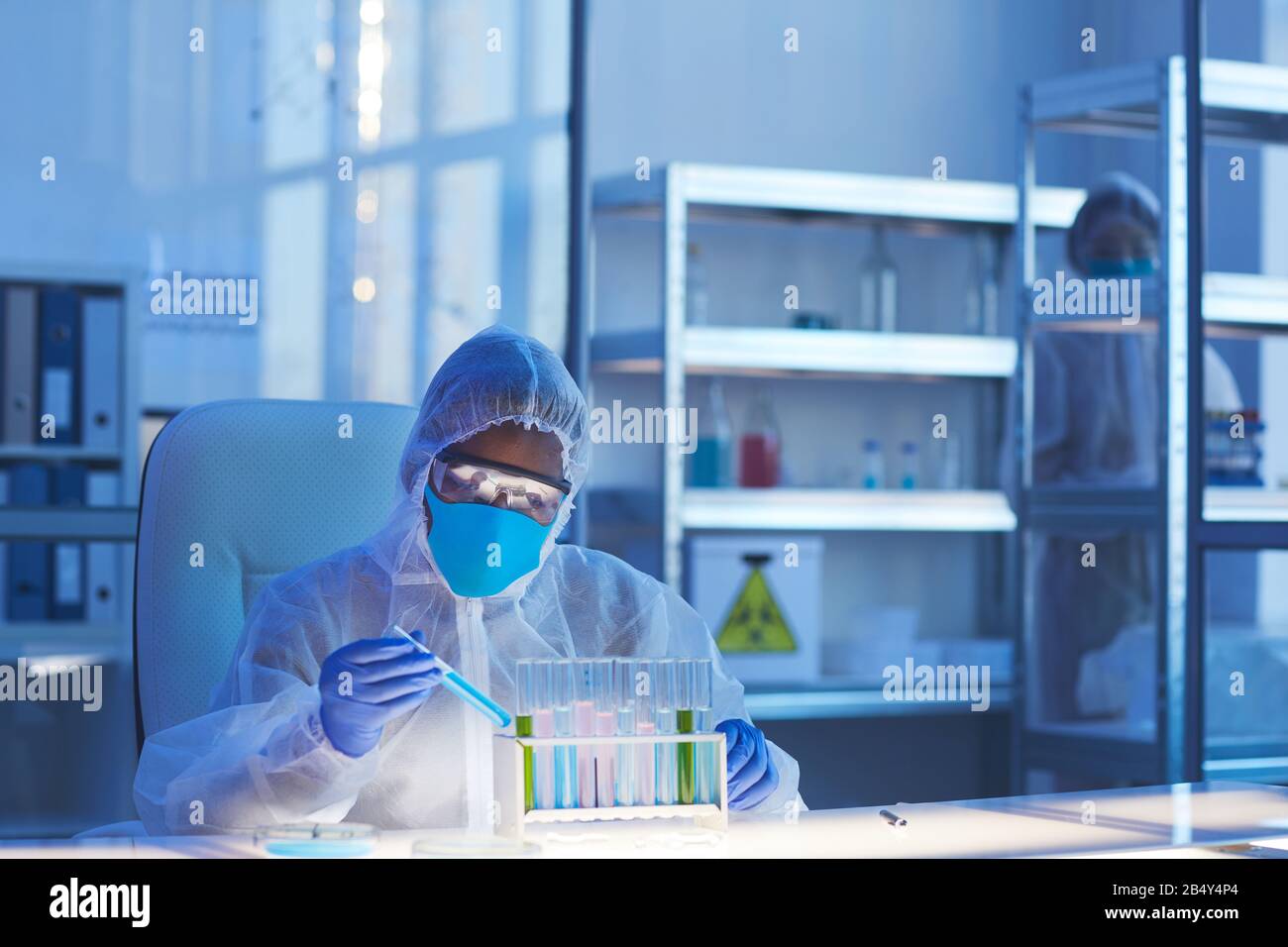 Horizontal shot of unrecognizable male medical scientist in protective clothes sitting at desk in modern laboratory holding test-tube with dangerous l Stock Photo