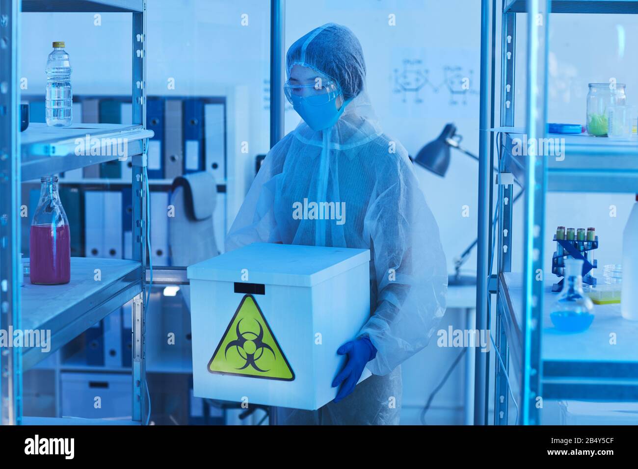 Horizontal medium long shot unrecognizable female laboratory worker in modern protective uniform holding box with biohazards Stock Photo