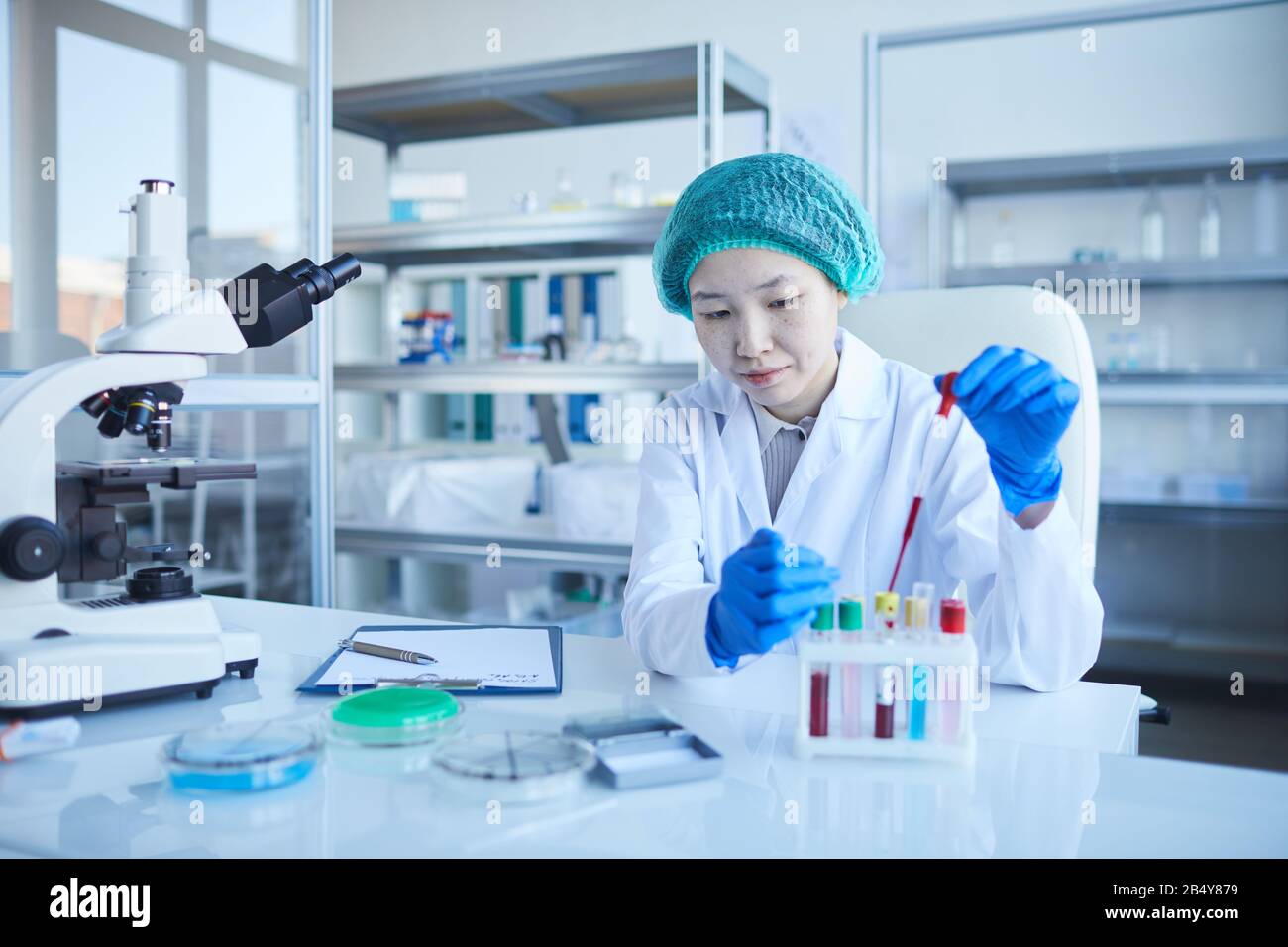 Horizontal portrait of concentrated young woman wearing lab coat, protective gloves and hat doing medical tests, copy space Stock Photo