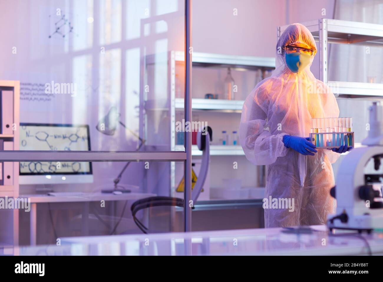 Unrecognizable female chemist wearing disposable protective uniform holding test-tubes with liquids looking at camera, horizontal medium long shot, co Stock Photo