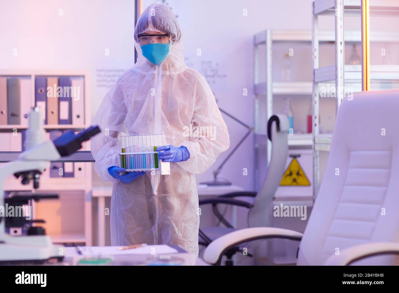 Unrecognizable female chemical industry worker wearing disposable protective uniform holding test-tubes with liquids, horizontal medium long shot, cop Stock Photo