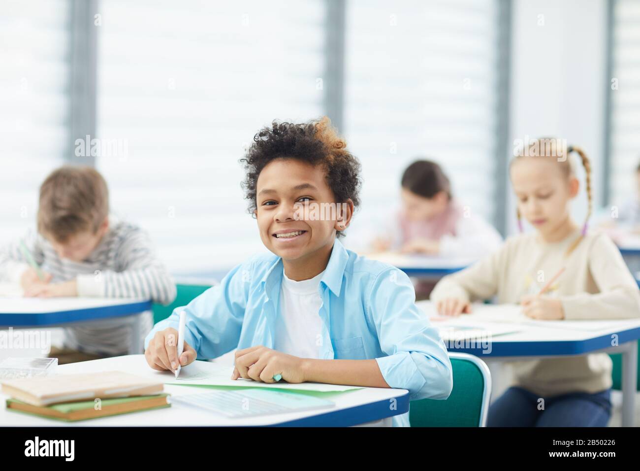 Horizontal medium close up portrait of happy mixed-race boy with kinky hair sitting at school desk looking at camera smiling, copy space Stock Photo