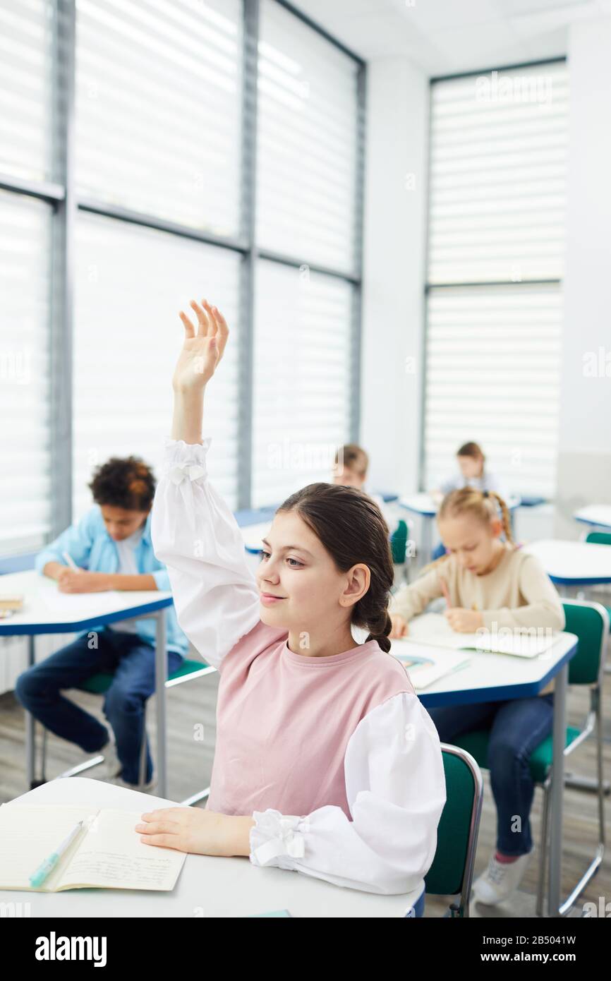 Vertical portrait of active smart twelve-year-old girl with dark hair sitting at school desk raising her hand in class, copy space Stock Photo
