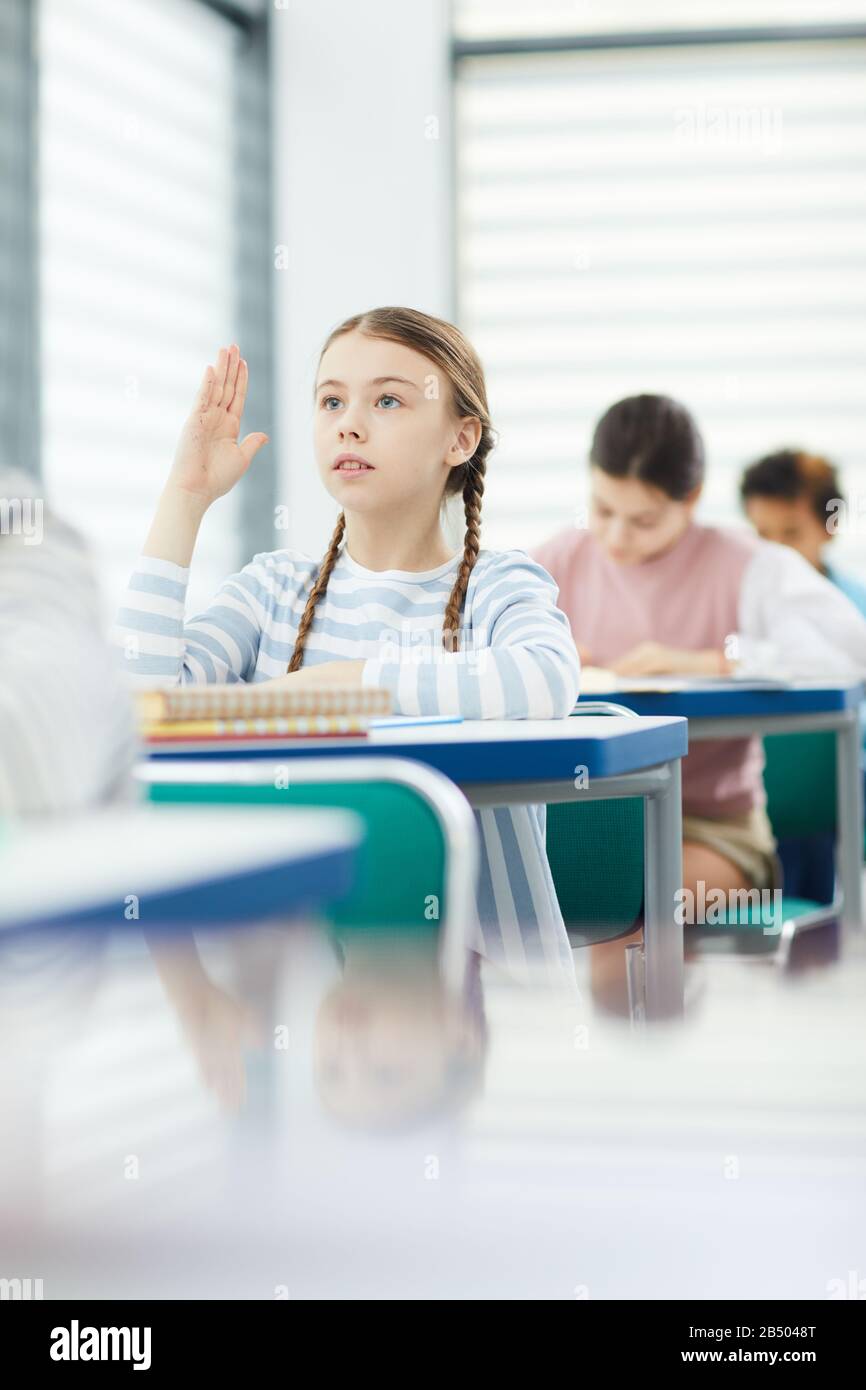 Vertical portrait of young schoolgirl sitting and desk raising hand and ...