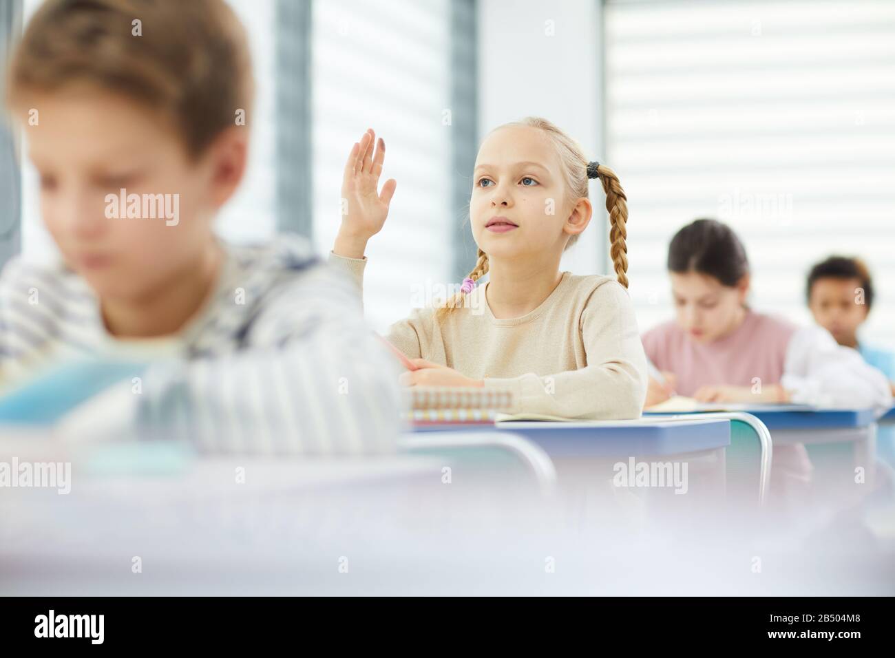 Horizontal portrait of pensive girl sitting at school desk raising her ...