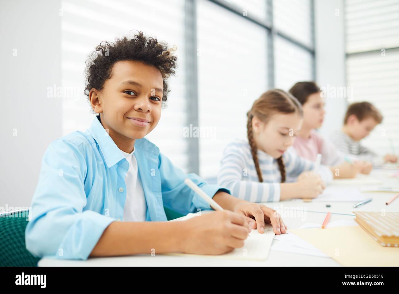 Portrait of cheerful African American boy sitting at school desk with ...
