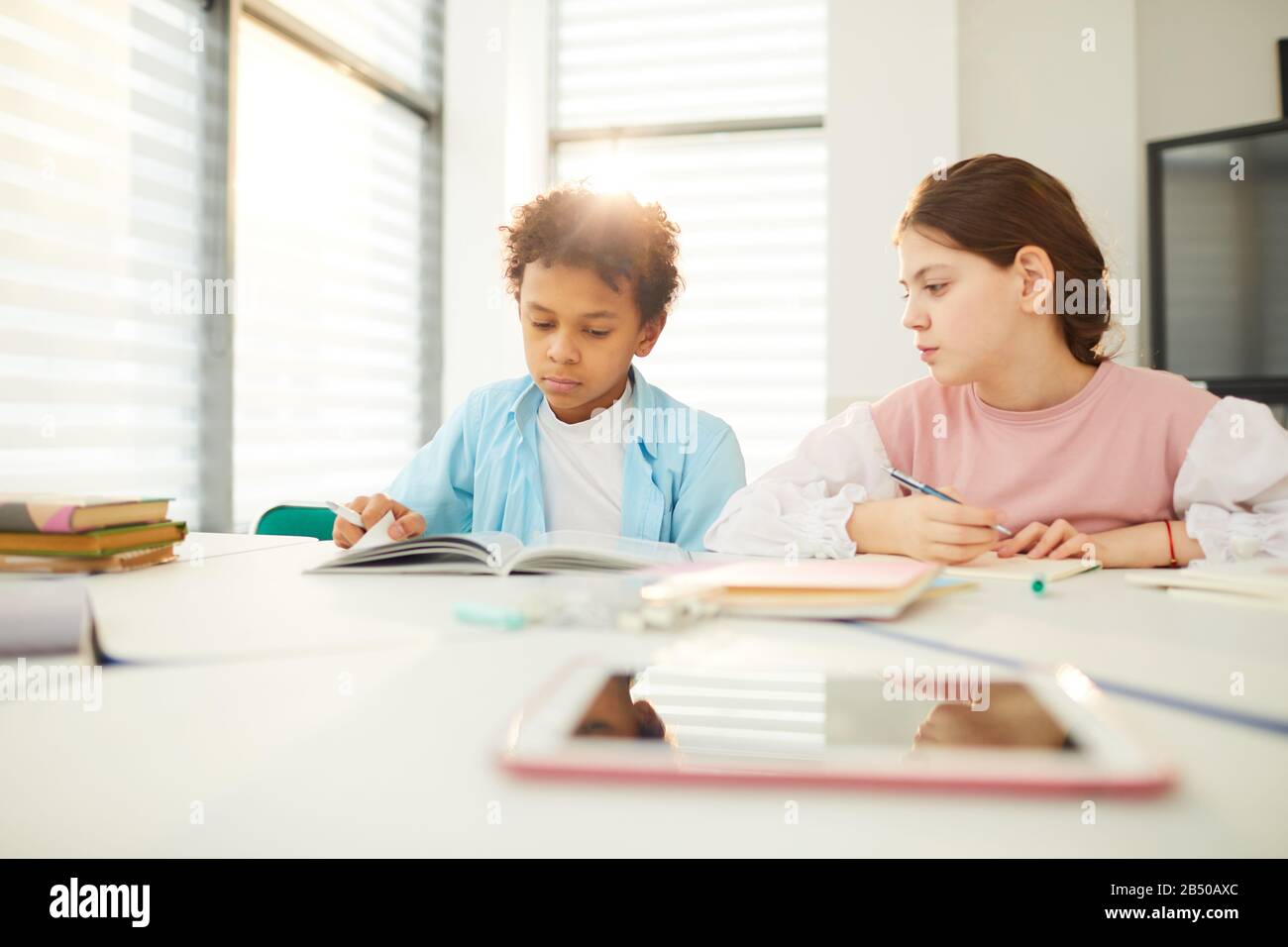 African American boy and Caucasian girl sitting together at school desk in modern classroom, girl looking at boy's textbook, copy space Stock Photo