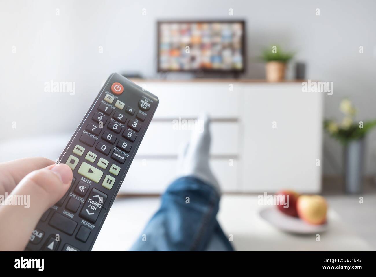 Man watching TV, lying on sofa, legs on table. Person holding remote control in living room Stock Photo