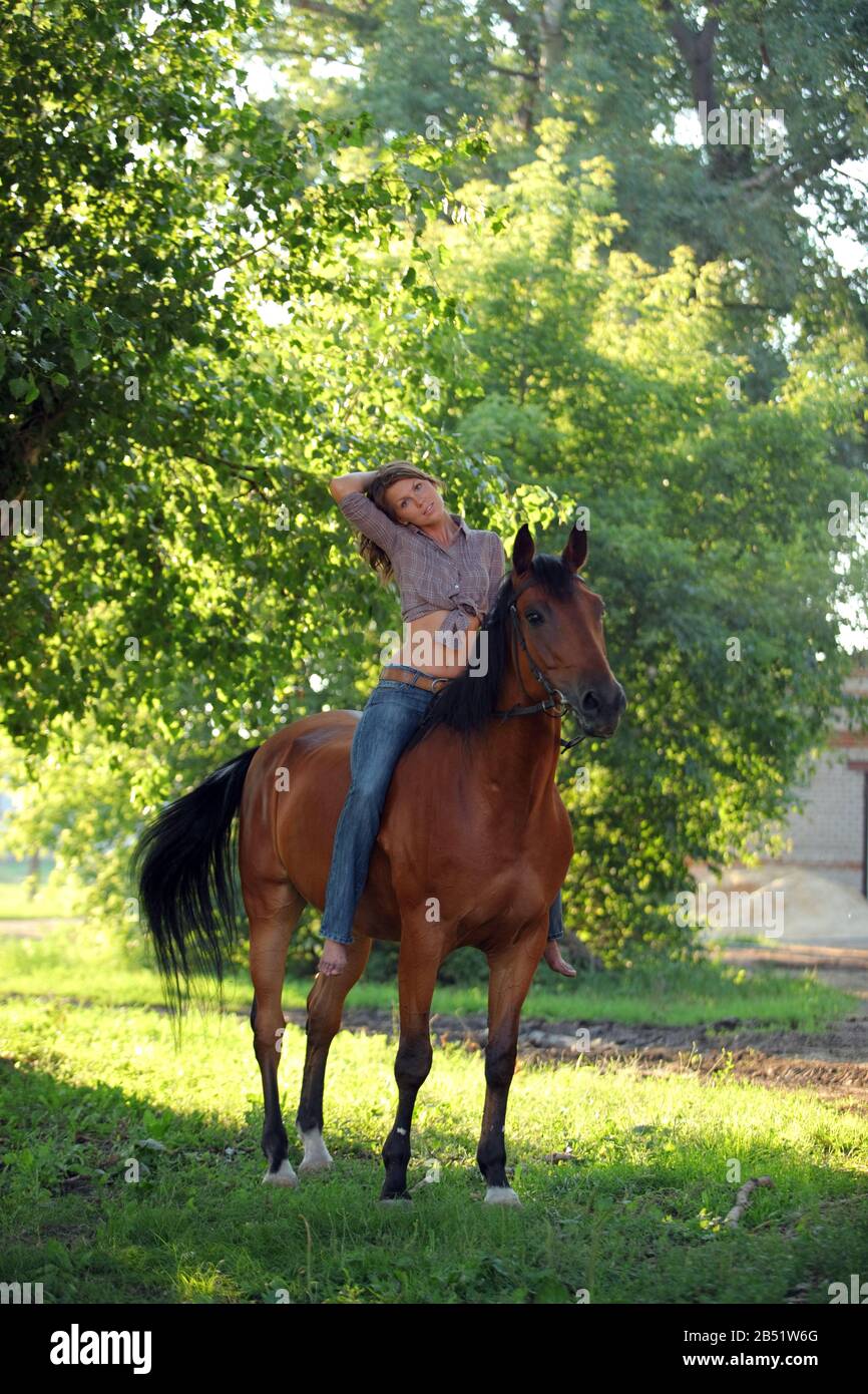Beautiful country woman walks with horse in woods glade at sunset Stock