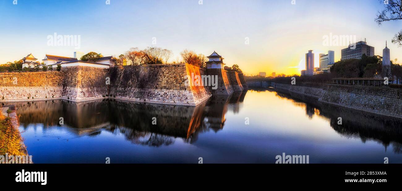 Wide moat around stone walls of historic palace, park and castle in Osaka city of Japan. Soft rising sun behind modern skyscrapers. Stock Photo