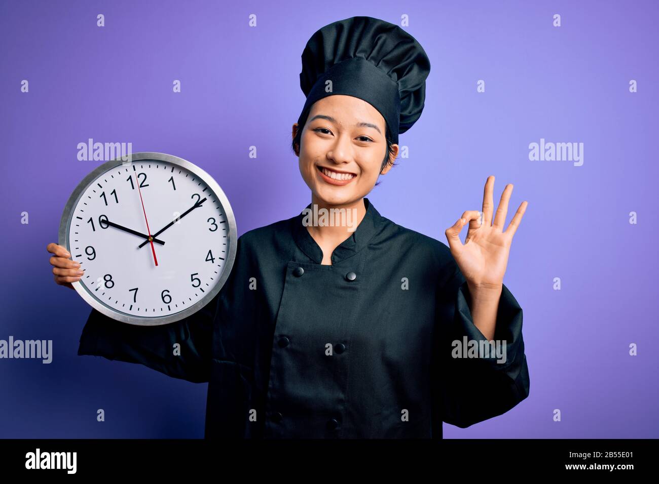 Young chinese chef woman wearing cooker uniform and hat holding clock doing countdown doing ok