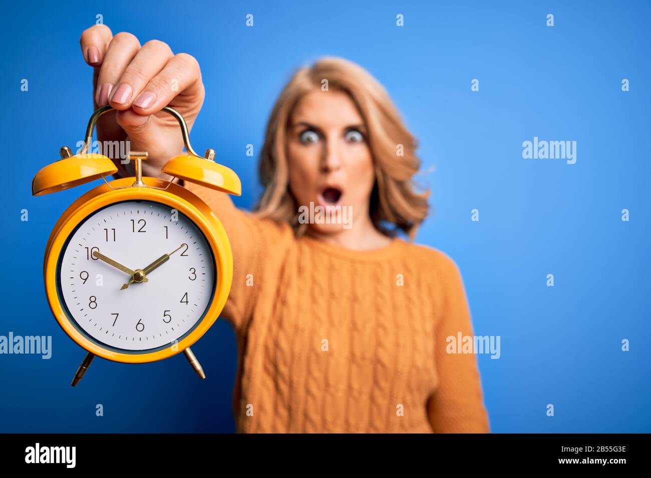 Middle age beautiful blonde woman holding vintage alarm clock over blue background scared in