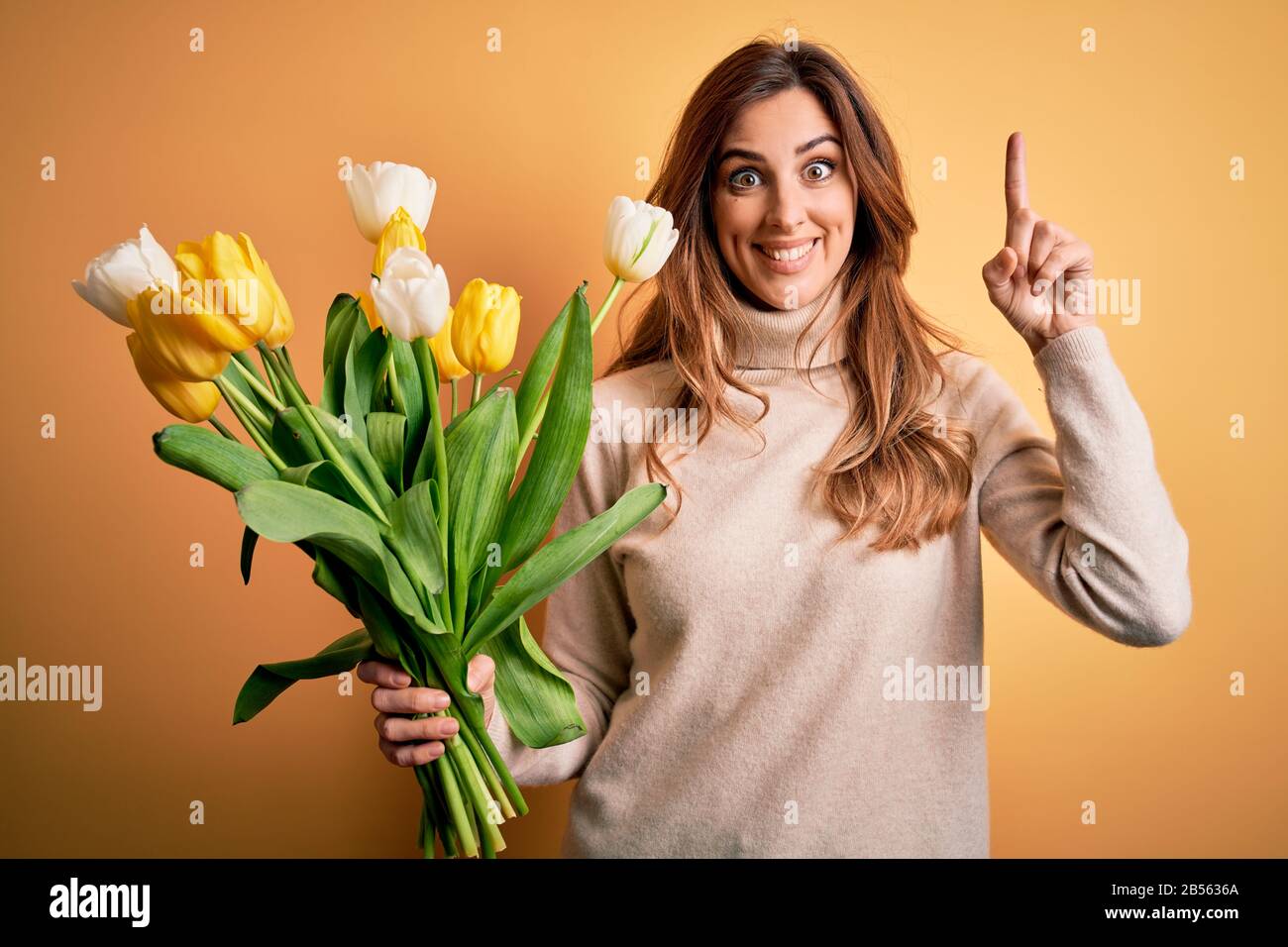 Young beautiful brunette woman holding bouquet of yellow tulips over isolated background smiling amazed and surprised and pointing up with fingers and Stock Photo