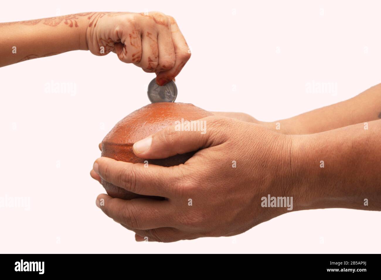 Hands Holding Indian penny bank made with mud clay and Child hand adding coin into the earthen pot or terracotta piggy bank on Isolated background - Stock Photo