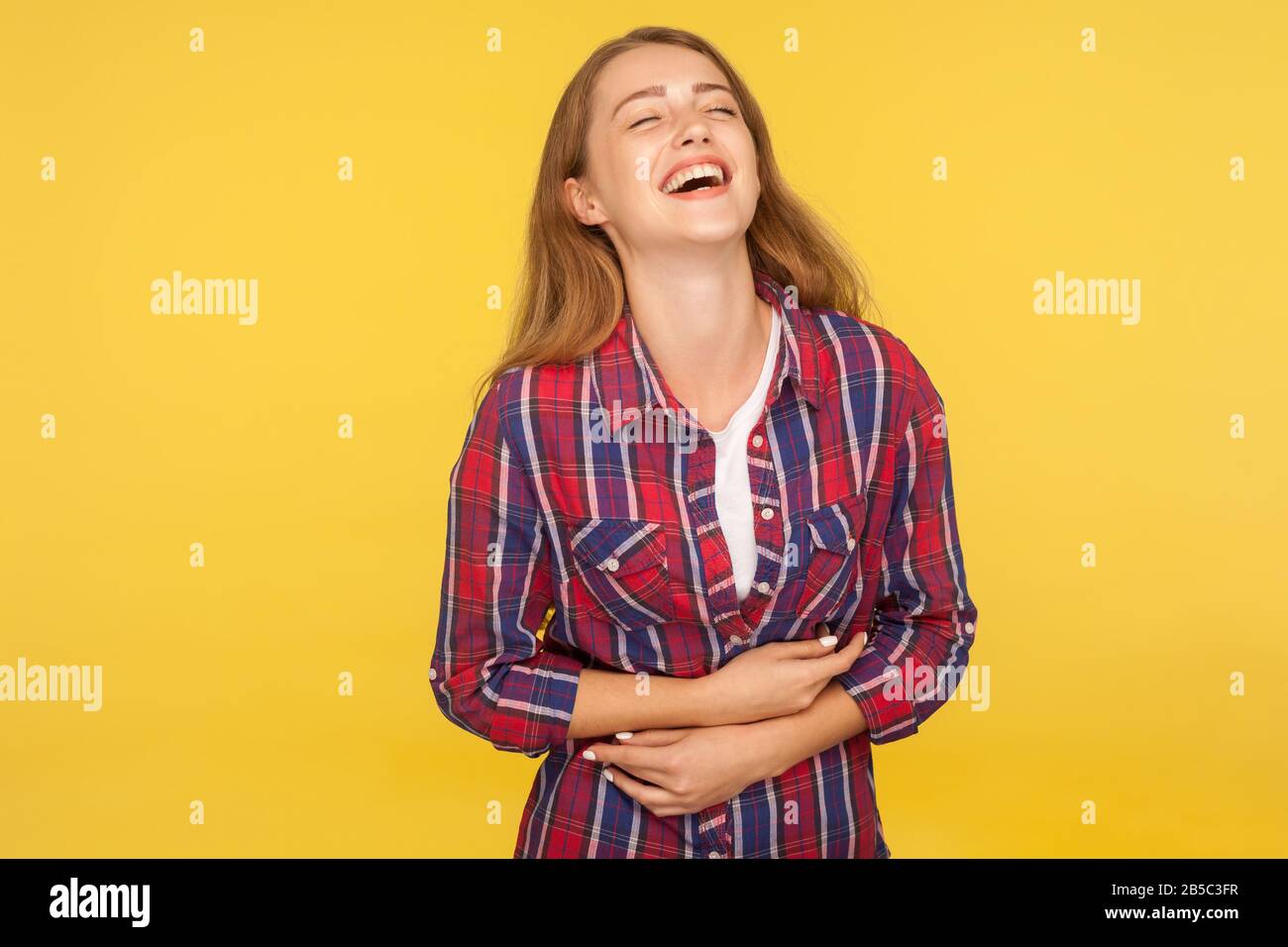 Portrait of carefree ginger girl in checkered shirt holding belly and laughing out loud after hearing hilarious joke, funny anecdote, being in good mo Stock Photo