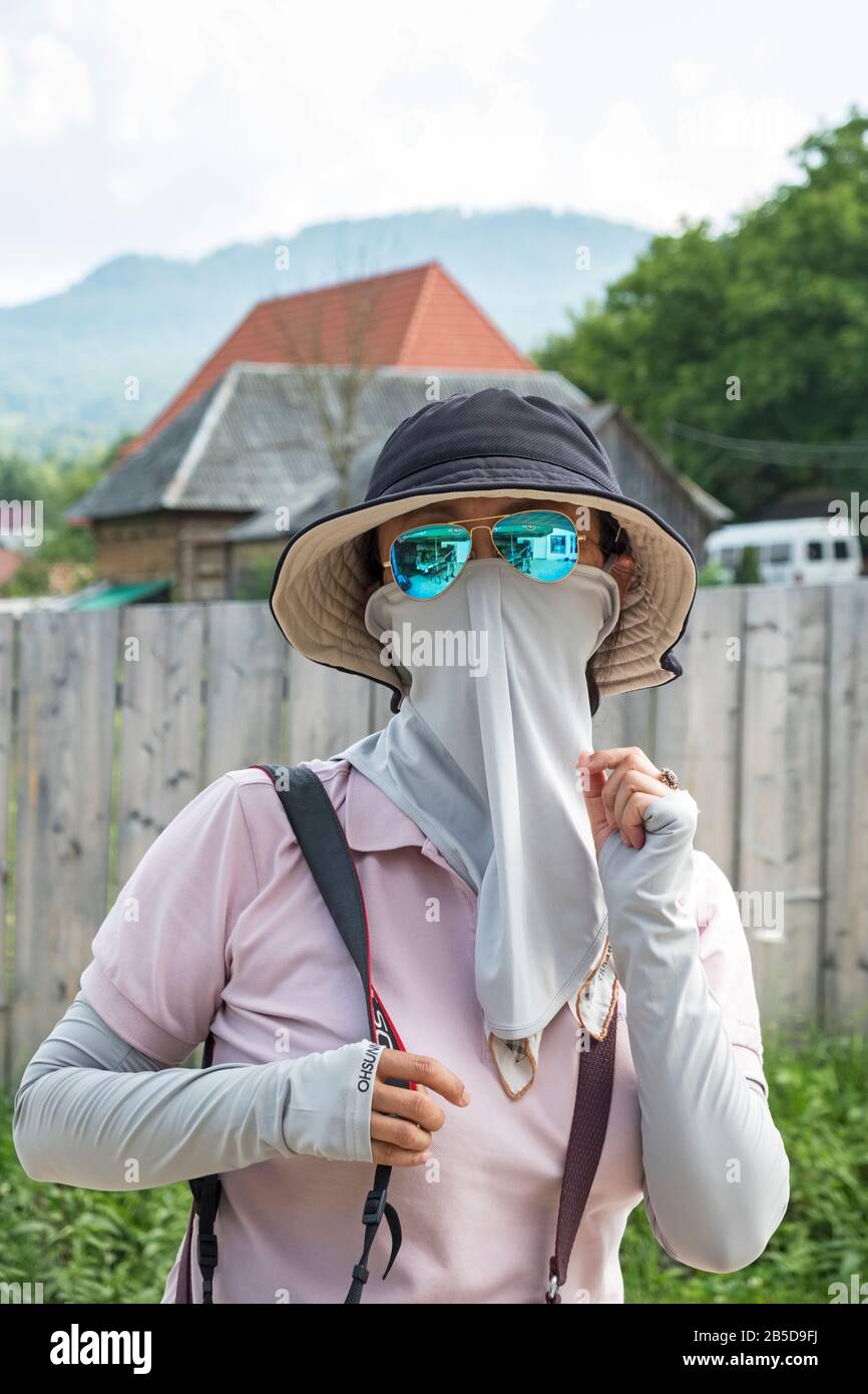 A Chinese woman tourist in Romania, wearing a straw hat, blue sunglasses, and a scarf or snood wrapped around her face as a protective facemask Stock Photo