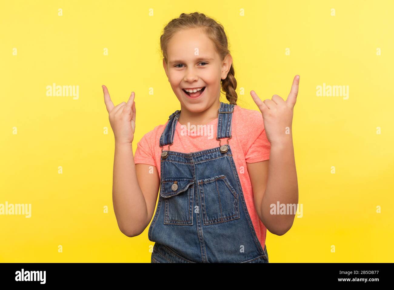 Portrait of overjoyed little girl with braid in denim overalls doing