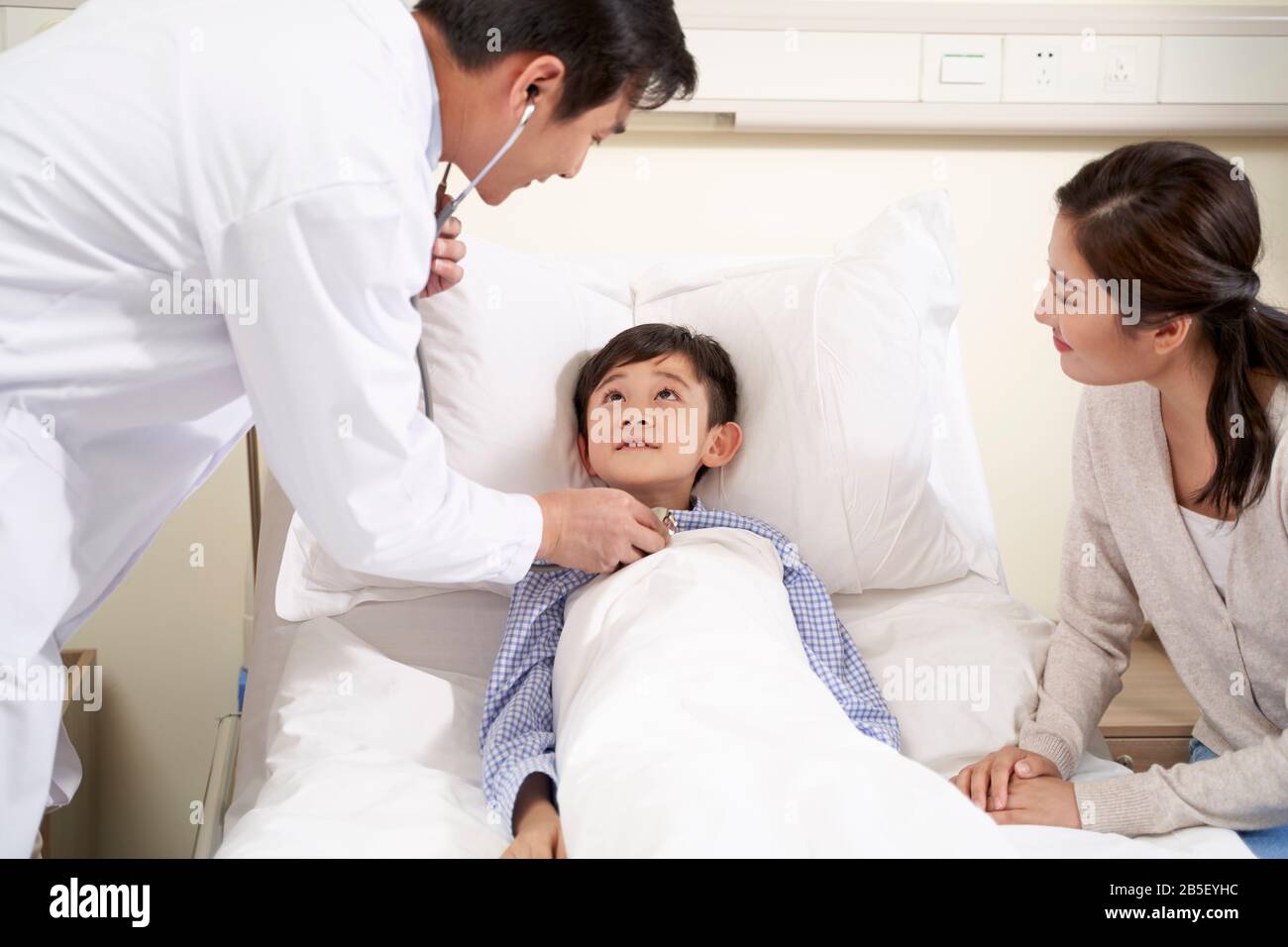 five year old asian kid lying in bed in hospital ward accompanied by mother and being examined by pediatrician using stethoscope Stock Photo