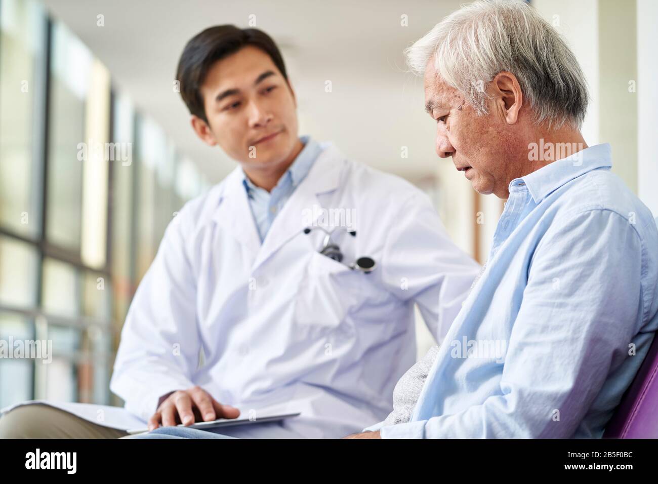 young asian doctor talking and explaining test result and diagnosis to demoralized elderly patient in hospital hallway Stock Photo
