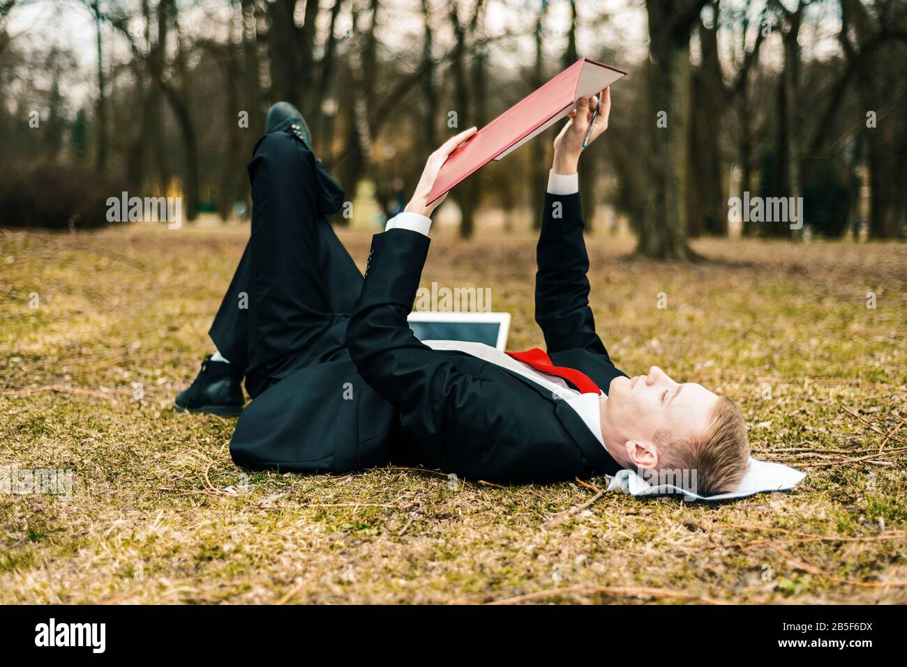 young guy lying in nature with laptop. businessman working remotely outdoors. freelancer reads documents. Stock Photo