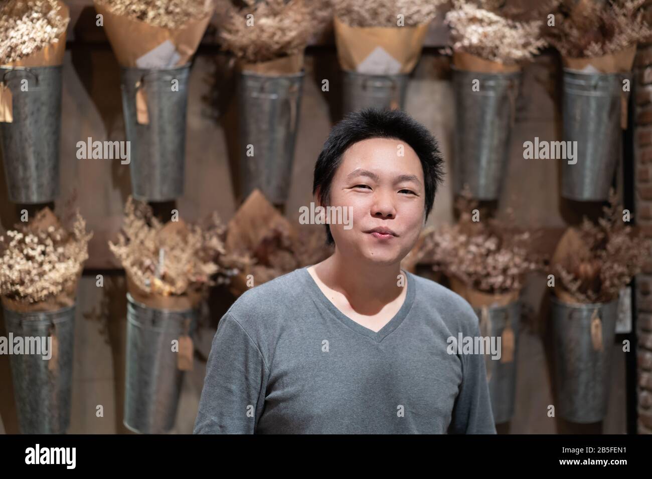 Happy Asian Adult man smiles to the camera in front of vertical dried flower in aluminum pot on the wall. Stock Photo