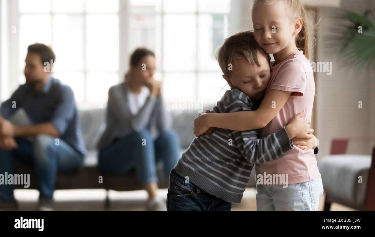 Stressed little girl embracing smaller brother, overcoming parents argue divorce. Stock Photo